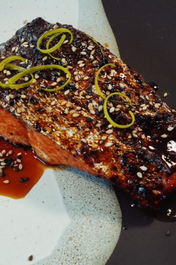 A grilled salmon fillet topped with sesame seeds and sliced green onions, served on a plate with a dark soy-based sauce.