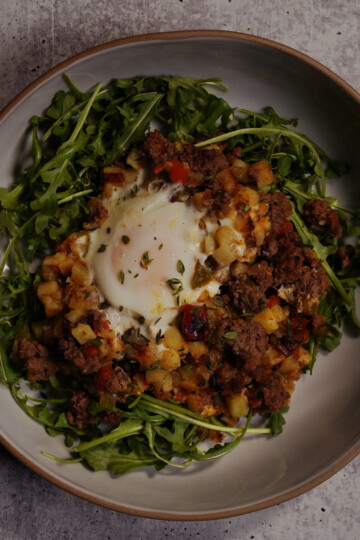 A bowl of arugula topped with a hash of diced potatoes, ground meat, vegetables, and a baked egg in the center, served on a gray textured surface.