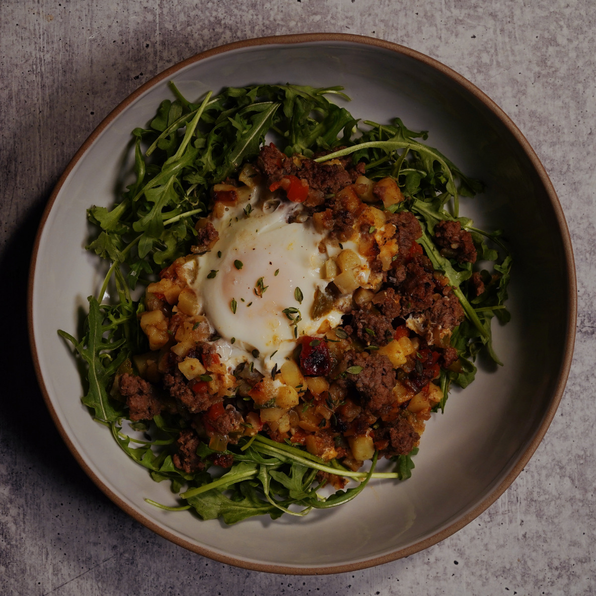 A bowl of arugula topped with a hash of diced potatoes, ground meat, vegetables, and a baked egg in the center, served on a gray textured surface.