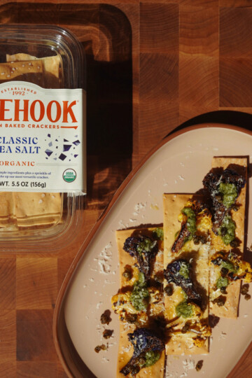 A container of Firehook Classic Sea Salt organic crackers sits next to a plate with several crackers topped with roasted vegetables, green sauce, grated cheese, and capers on a wooden surface.
