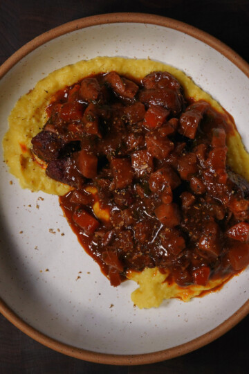 A plate of creamy yellow polenta topped with a rich, chunky beef stew in a brown sauce with visible pieces of carrots, served on a white plate with a brown rim.