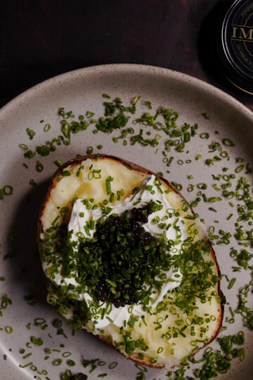 A baked potato topped with sour cream, black caviar, and finely chopped chives sits on a beige plate. A tin labeled Imperia caviar is visible in the corner of the image.