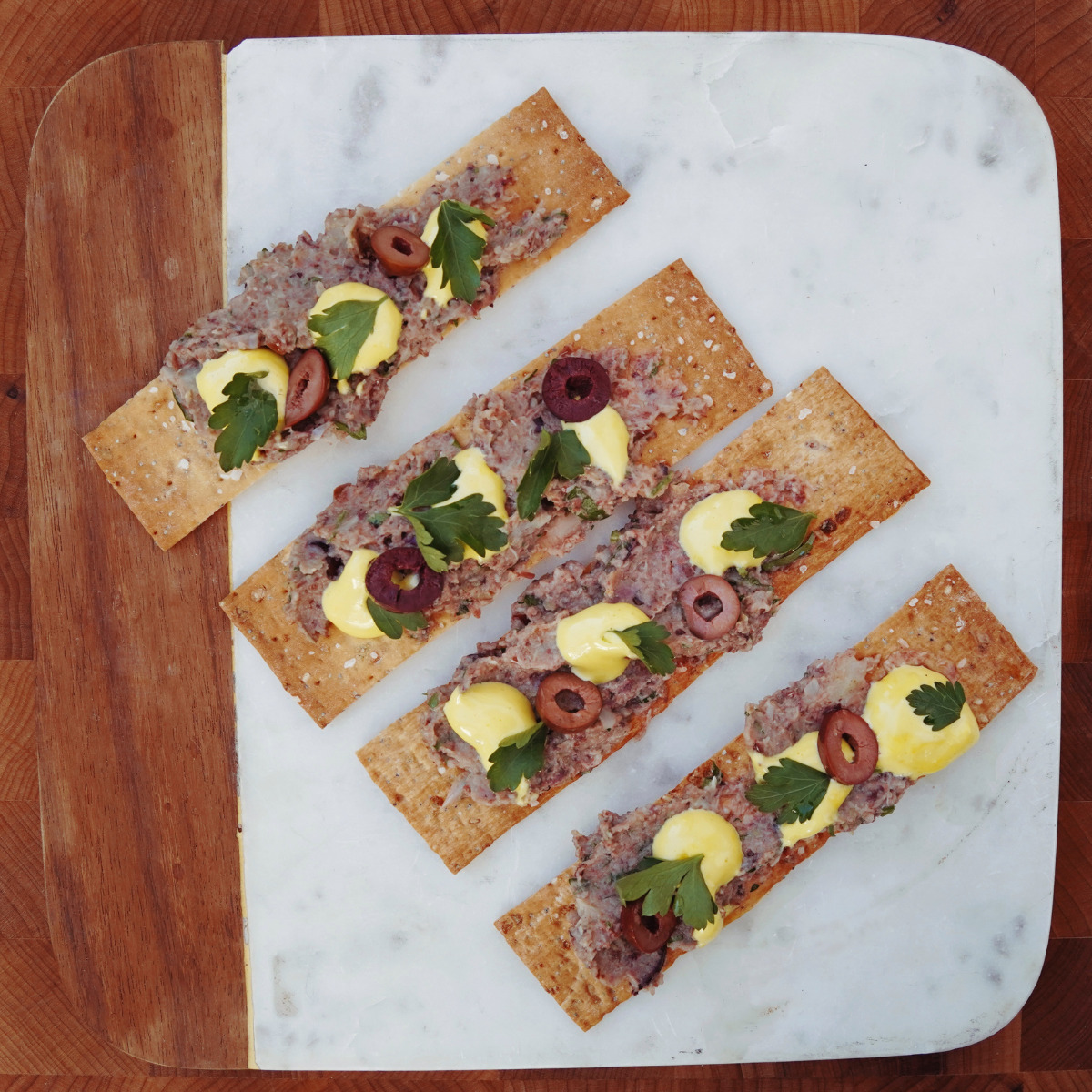 Four rectangular crispbread pieces topped with a spread, sliced olives, dollops of yellow sauce, and fresh parsley are arranged neatly on a marble and wood serving board.