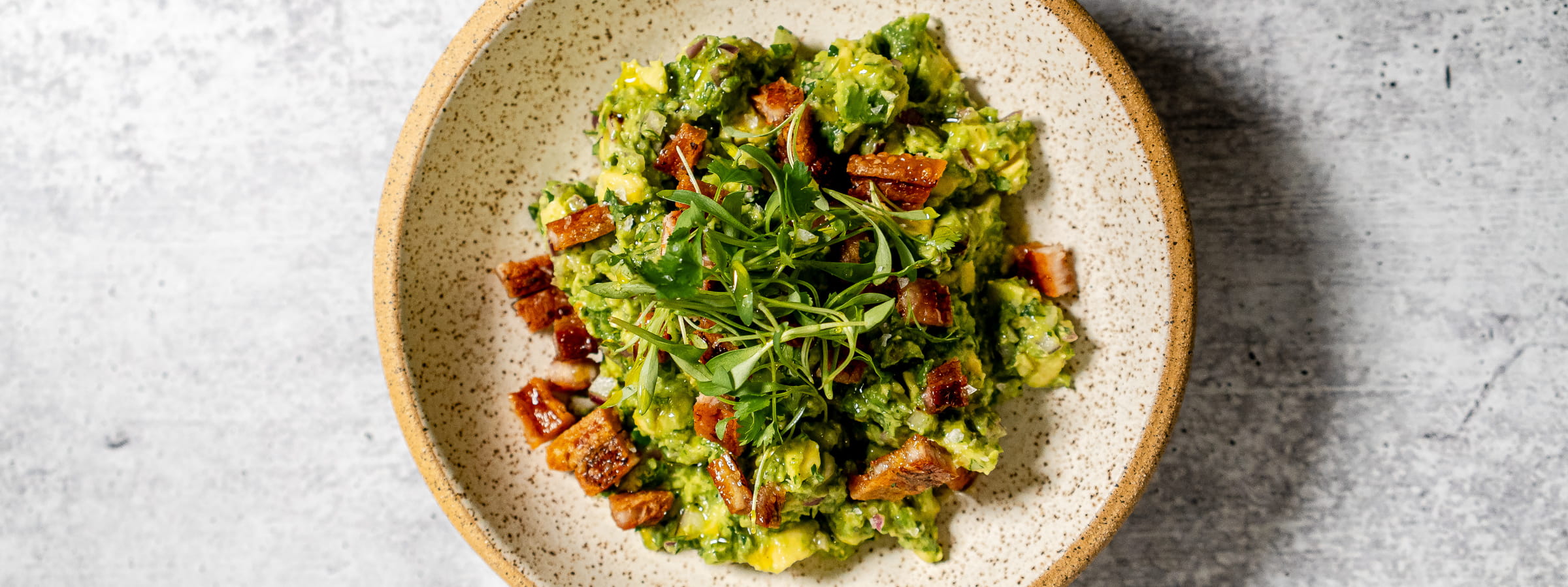 A speckled ceramic bowl filled with chunky avocado salad, topped with crispy browned cubes and fresh green herbs, sits on a light gray textured surface.