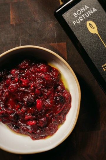 A bowl of chunky red cranberry sauce sits on a dark wooden surface next to a bottle of Bona Furtuna extra virgin olive oil.