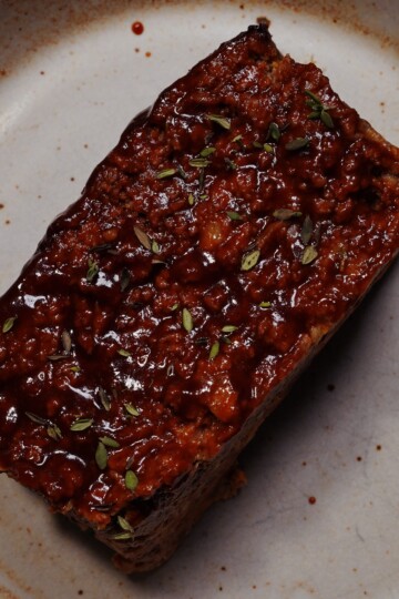 A rectangular slice of glazed meatloaf sits on a beige ceramic plate, garnished with small green herb leaves.