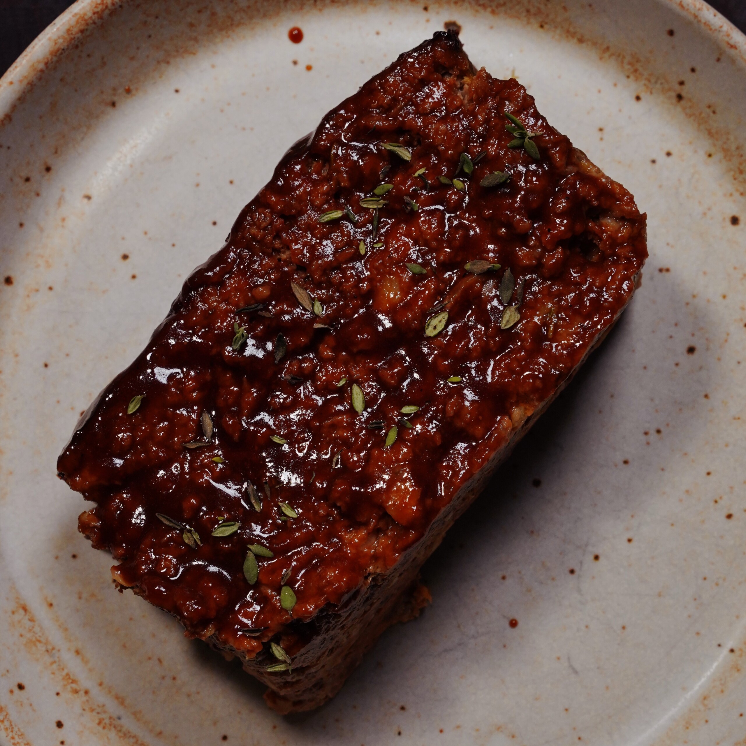 A rectangular slice of glazed meatloaf sits on a beige ceramic plate, garnished with small green herb leaves.