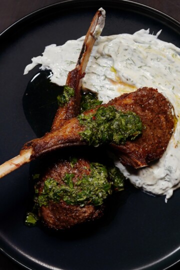 Two grilled lamb chops topped with green chimichurri sauce, served on a black plate with a generous smear of creamy white tzatziki sauce.