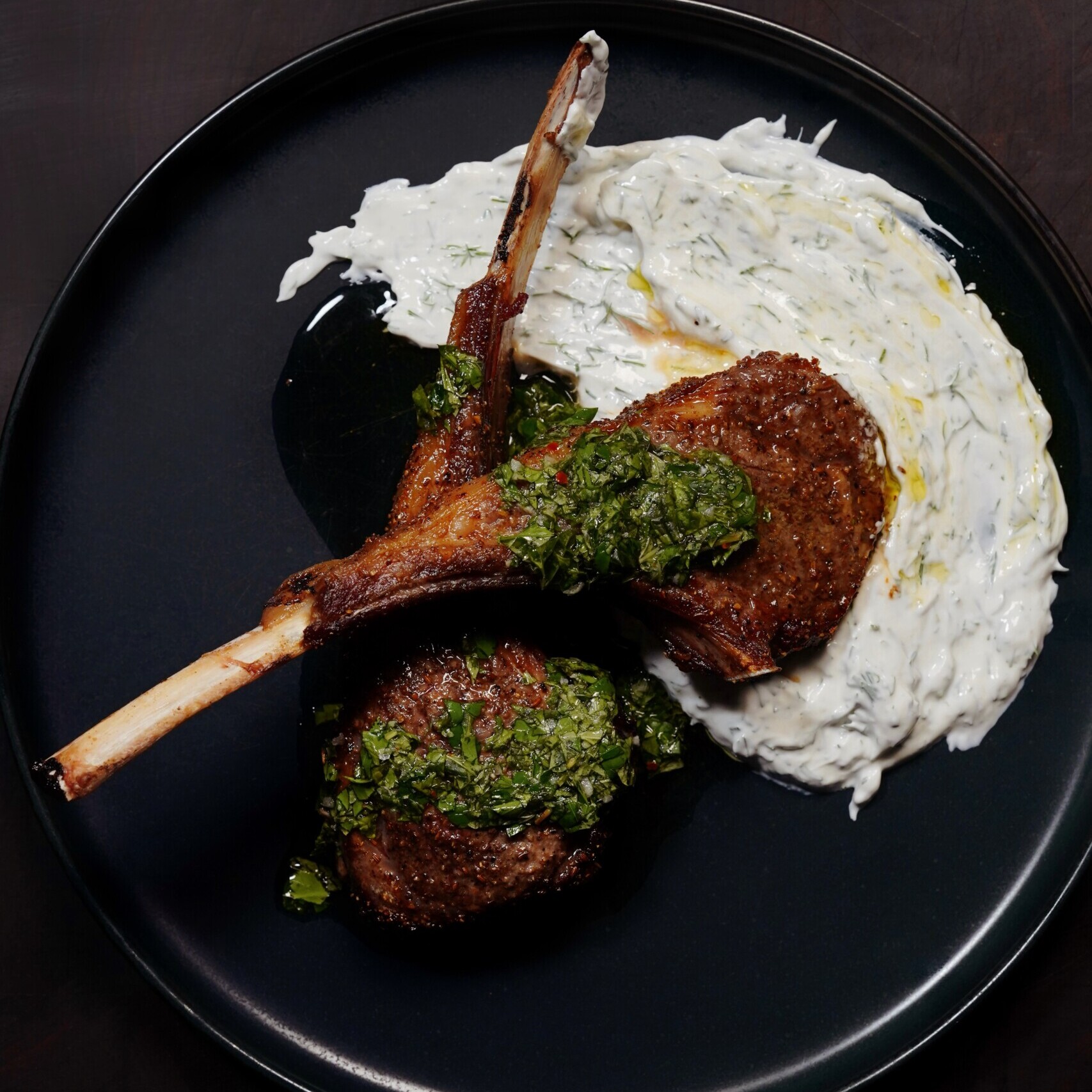 Two grilled lamb chops topped with green chimichurri sauce, served on a black plate with a generous smear of creamy white tzatziki sauce.