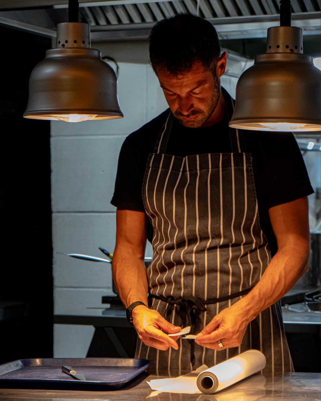 A chef wearing a striped apron stands under two hanging lights, carefully preparing food on a counter with a tray and a roll of parchment paper. The kitchen background is dimly lit.