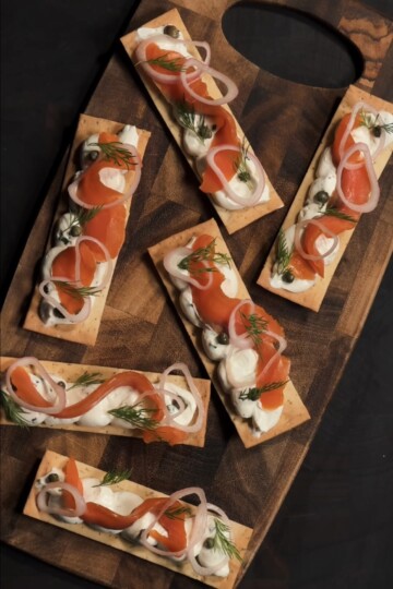Rectangular crackers topped with cream, smoked salmon, capers, sliced red onions, and fresh dill, arranged neatly on a wooden serving board against a dark background.