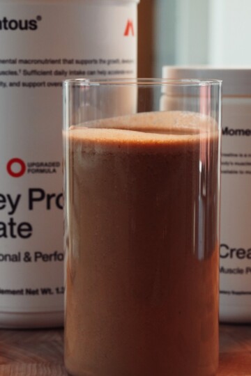 A glass of chocolate protein shake sits in front of containers labeled Momentous Whey Protein Isolate and Momentous Creatine on a wooden surface.