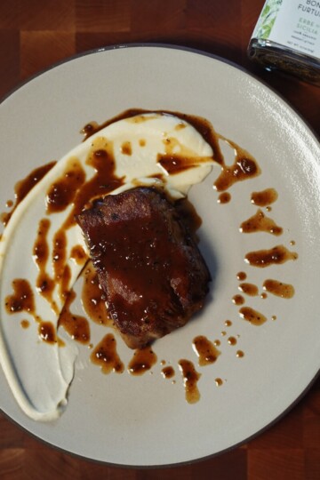 A plated dish featuring a piece of glazed meat with a creamy white sauce artistically swirled on a beige plate, drizzled with brown sauce, with a jar of seasoning partially visible in the corner.