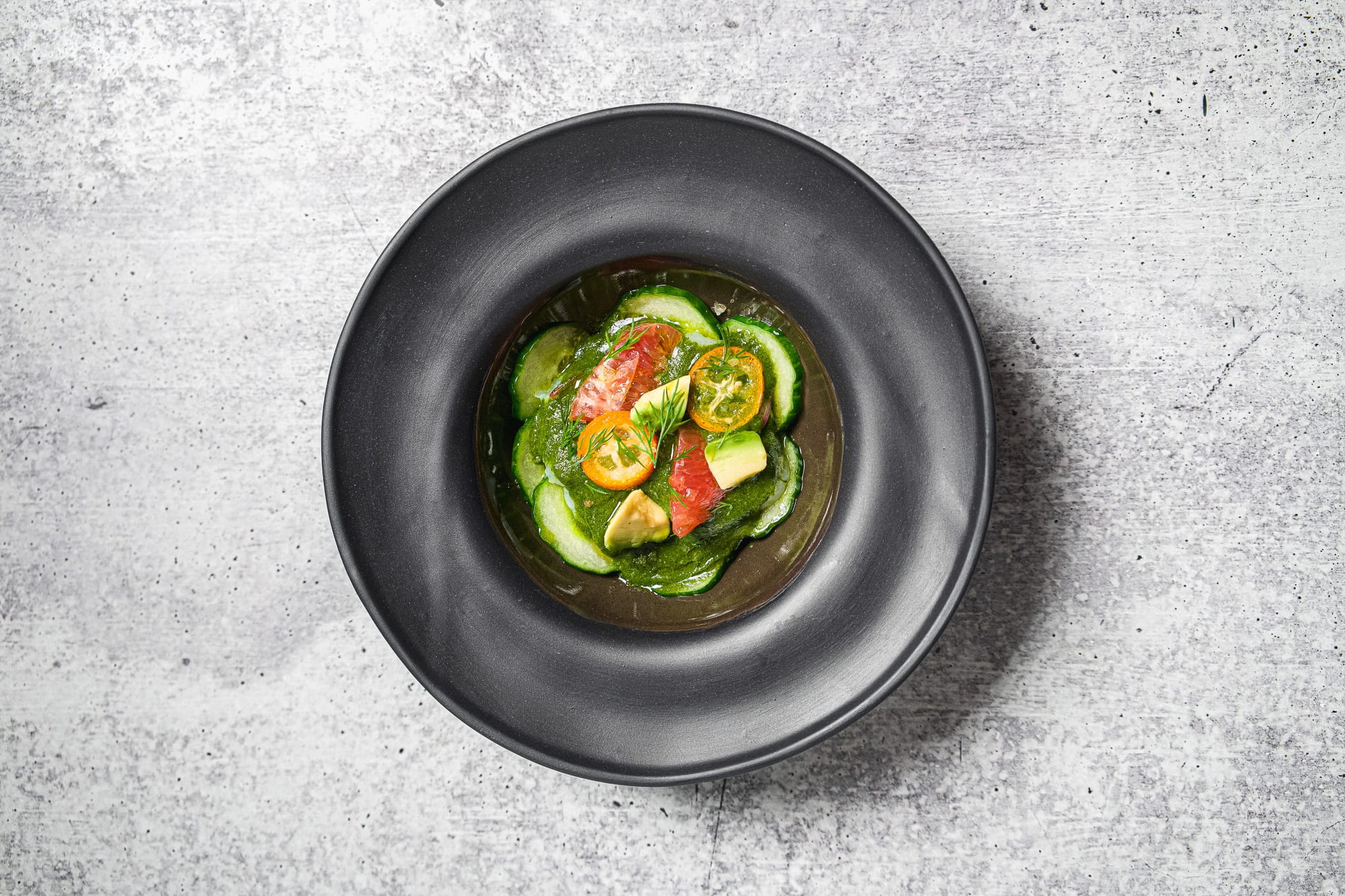 A black bowl filled with a fresh salad featuring slices of cucumber, grapefruit, and small citrus fruits, all arranged in a light dressing, against a light gray textured background.