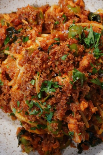 A close-up of a plate of spaghetti topped with a chunky tomato sauce, green olives, fresh chopped parsley, and a generous layer of golden brown breadcrumbs. The dish is served on a speckled ceramic plate.