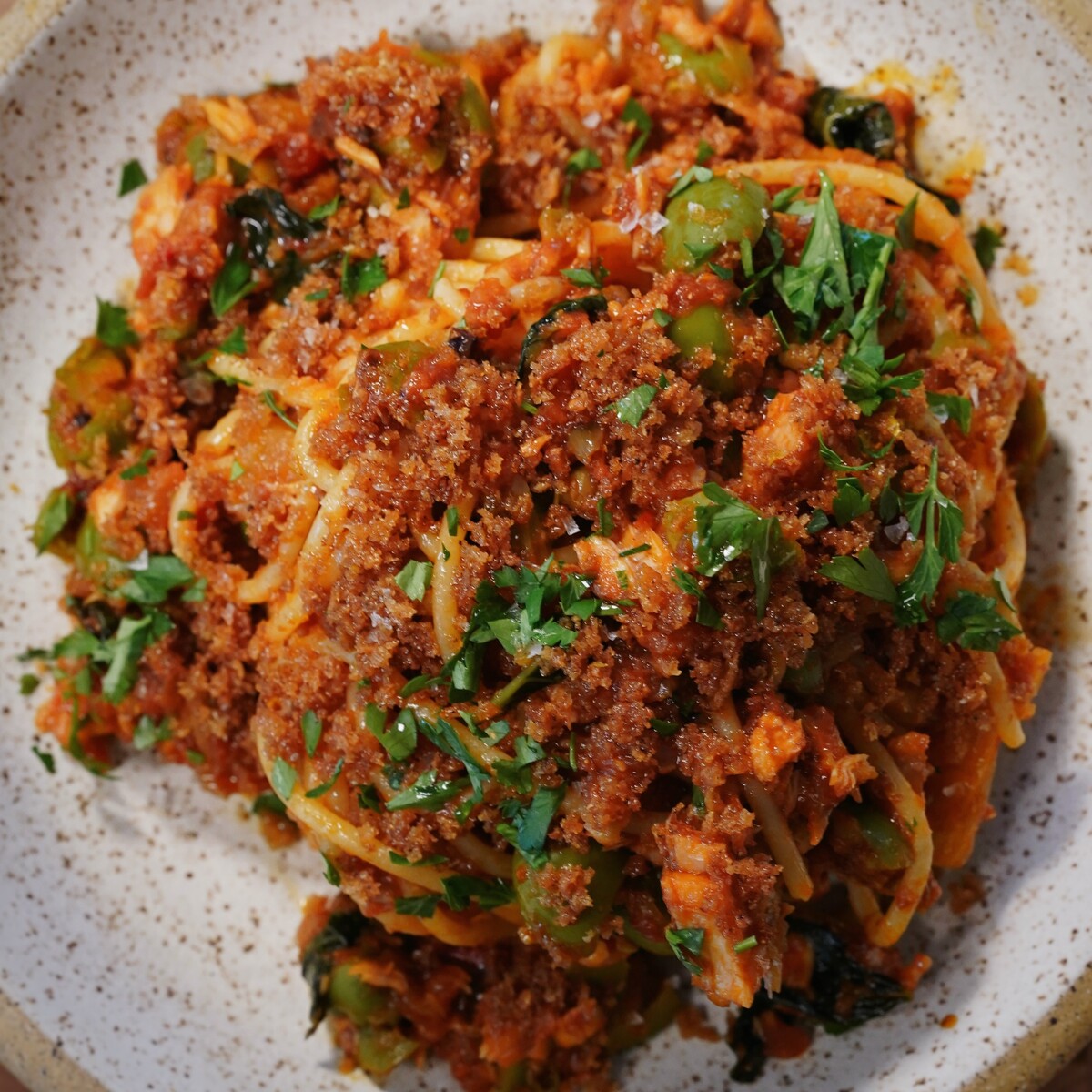 A close-up of a plate of spaghetti topped with a chunky tomato sauce, green olives, fresh chopped parsley, and a generous layer of golden brown breadcrumbs. The dish is served on a speckled ceramic plate.