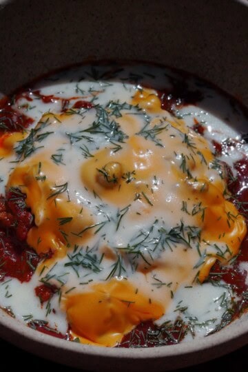 A bowl of stew topped with melted cheese, a creamy white sauce, and garnished with chopped herbs, served in a ceramic dish. The sauce and cheese are spread over a rich, red tomato-based mixture.