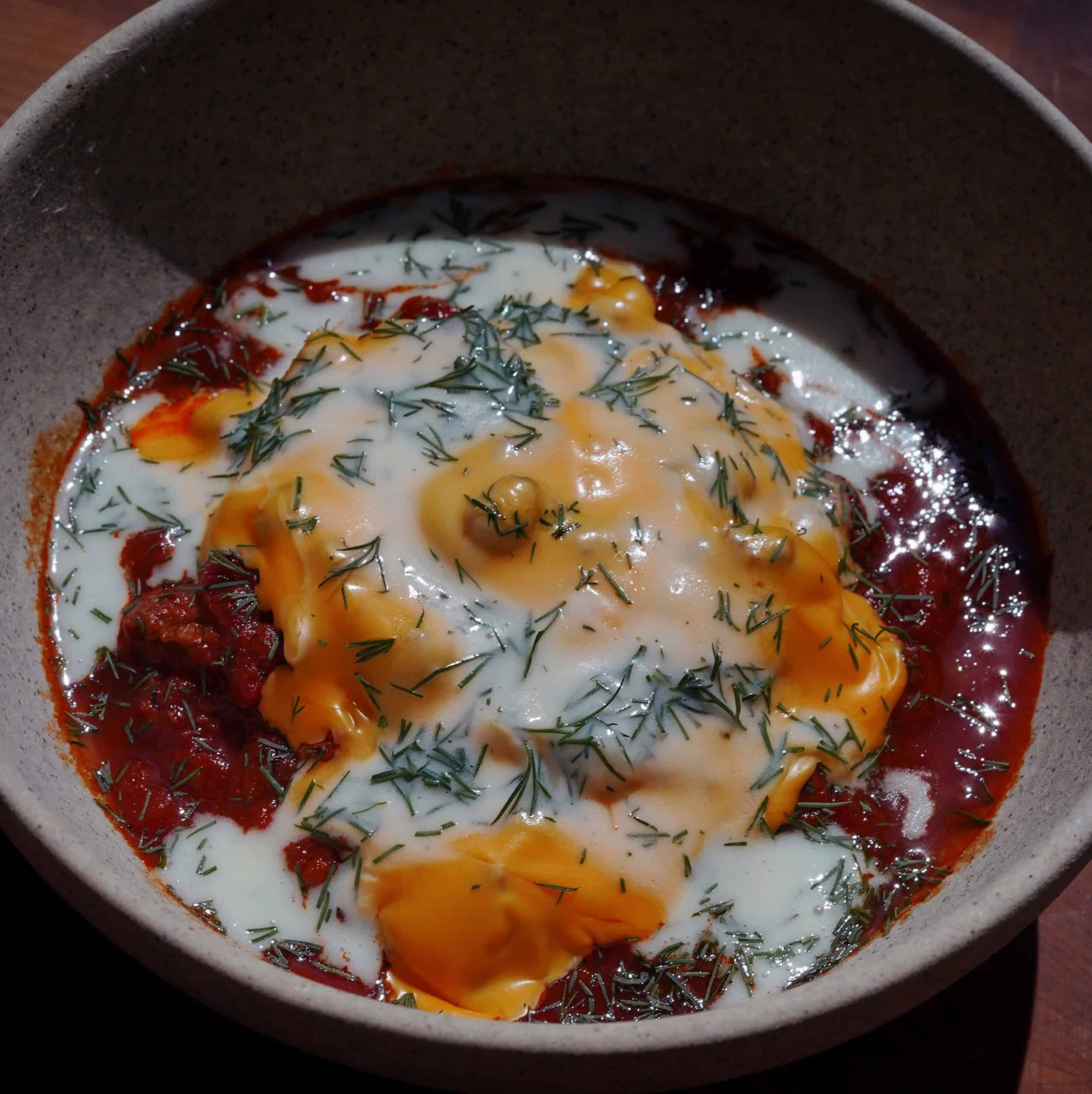 A bowl of stew topped with melted cheese, a creamy white sauce, and garnished with chopped herbs, served in a ceramic dish. The sauce and cheese are spread over a rich, red tomato-based mixture.