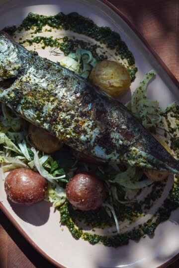 A whole roasted fish topped with green herbs sits on a platter with roasted red potatoes, sliced fennel, and greens, drizzled with herb sauce. The food is arranged on an oval dish on a wooden table.
