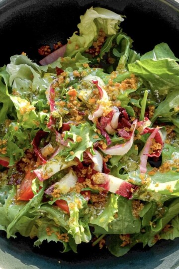 A bowl of fresh mixed greens salad with arugula, red onions, cherry tomatoes, and a sprinkling of golden crumbs, drizzled with a light dressing.