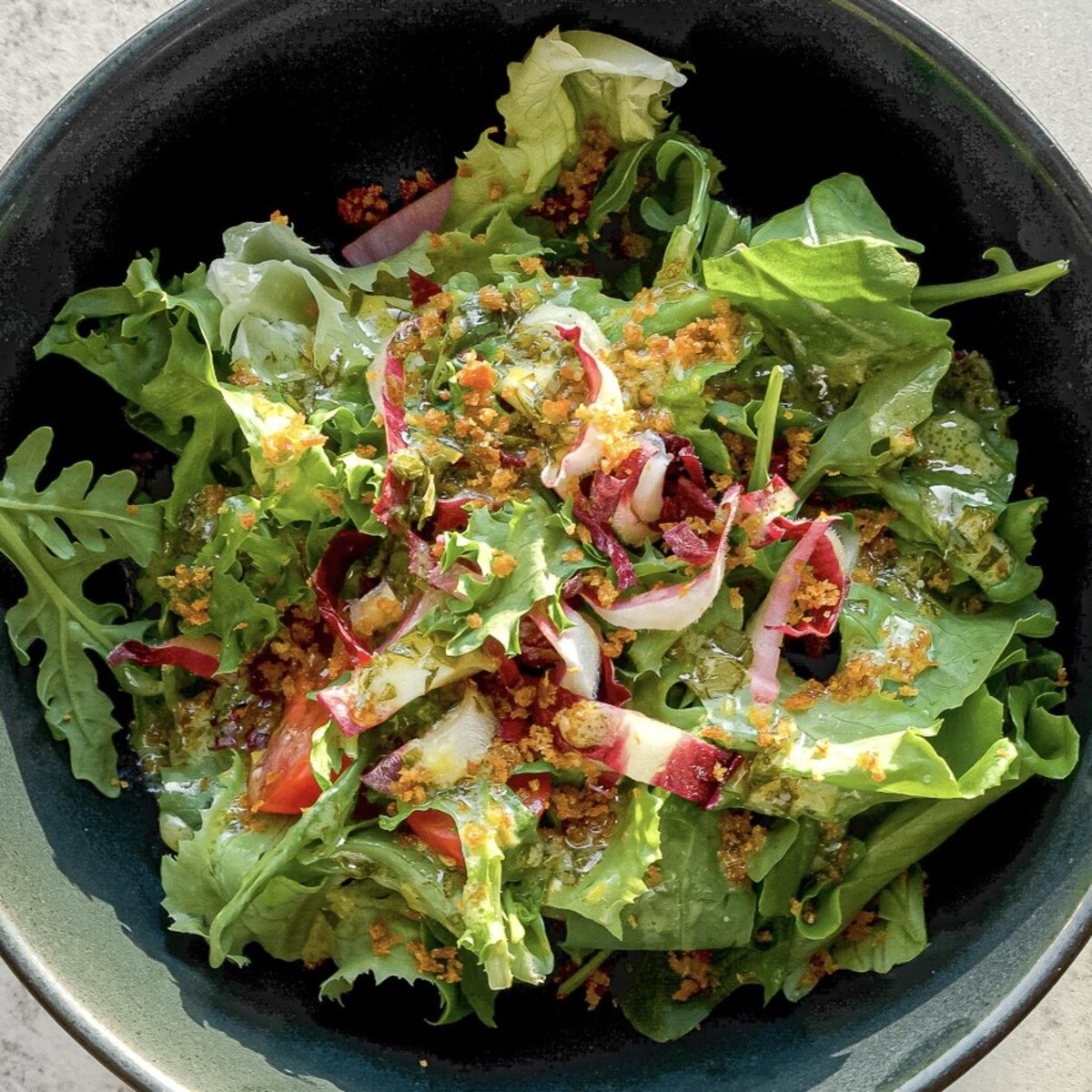 A bowl of fresh mixed greens salad with arugula, red onions, cherry tomatoes, and a sprinkling of golden crumbs, drizzled with a light dressing.
