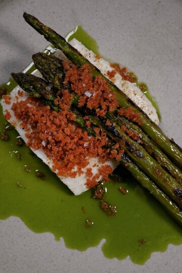 A beige plate with grilled asparagus spears on top of a piece of white fish, garnished with reddish-brown crumbs and surrounded by a green sauce.