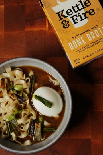 A bowl of noodle soup with scallions and a boiled egg sits on a wooden surface next to a carton of Kettle & Fire miso ginger beef bone broth.