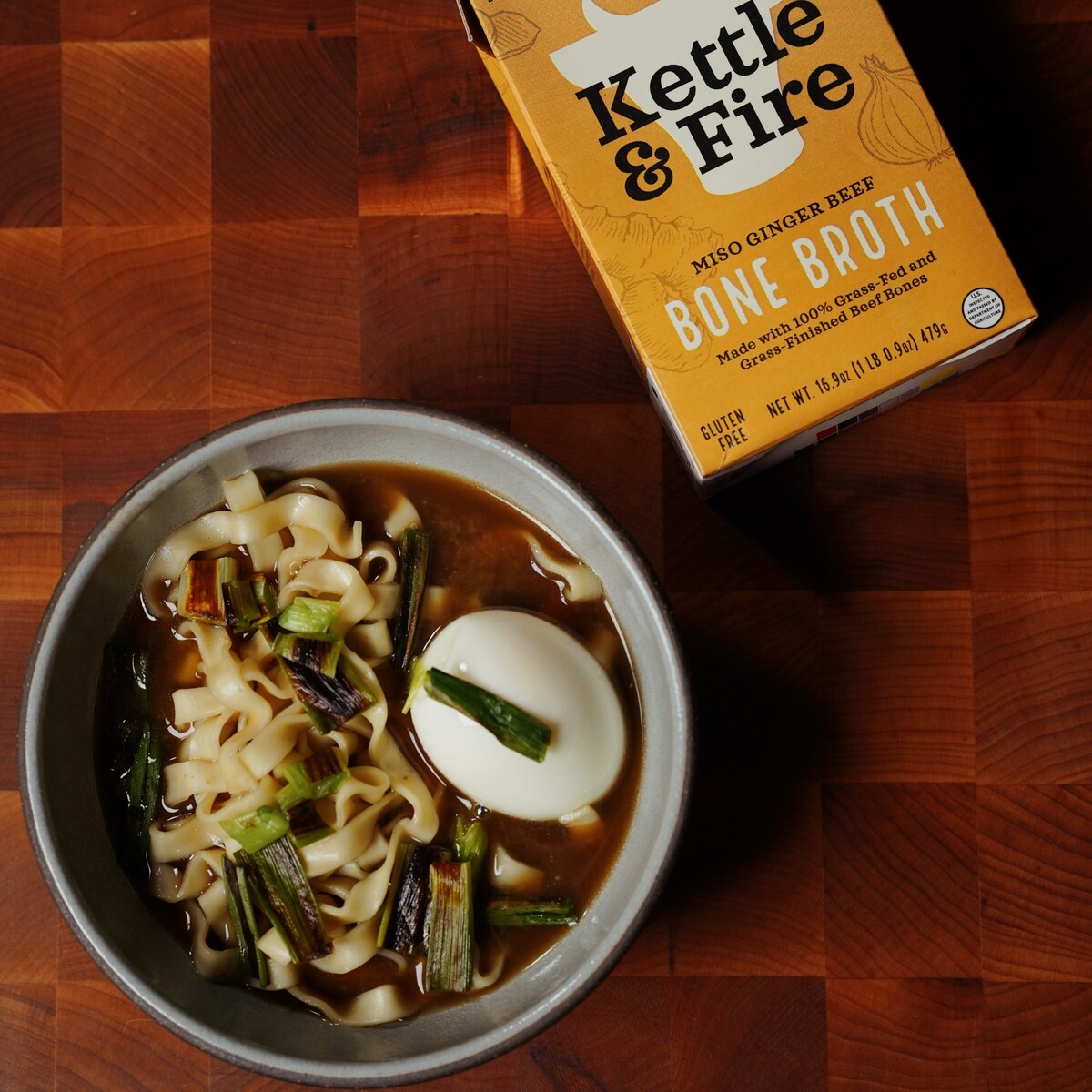 A bowl of noodle soup with scallions and a boiled egg sits on a wooden surface next to a carton of Kettle & Fire miso ginger beef bone broth.