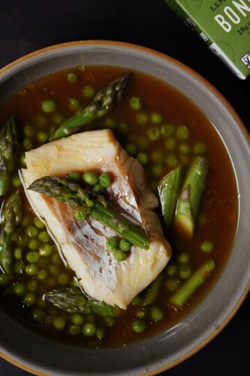 A bowl of fish fillet in broth is garnished with asparagus spears and green peas. A box labeled Bone Broth is partially visible in the upper right corner on a dark surface.