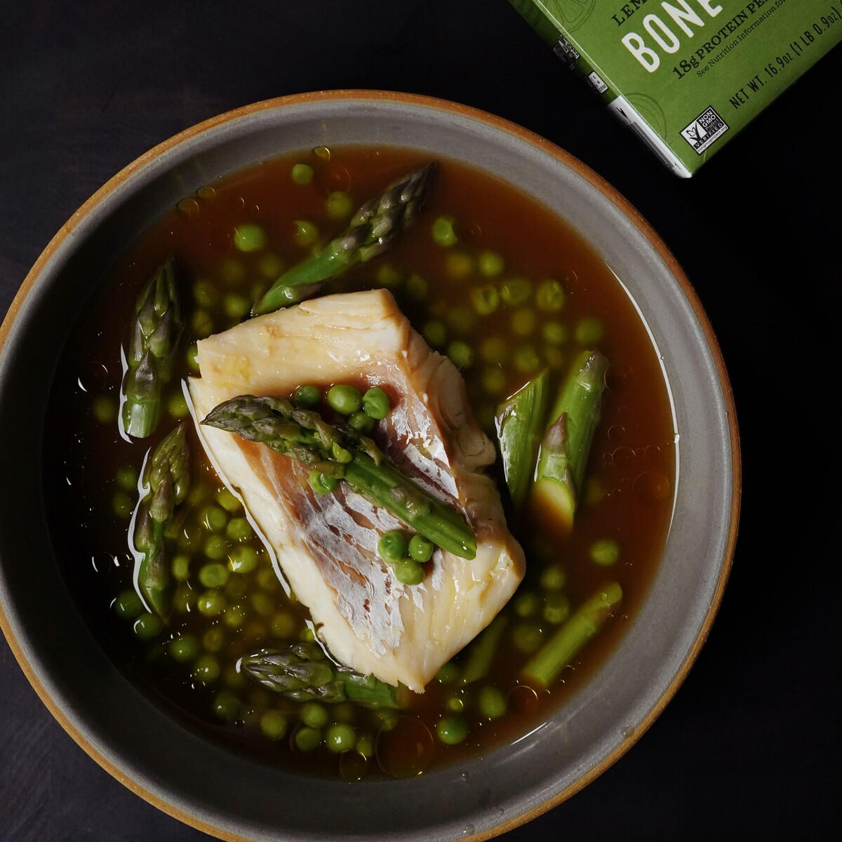 A bowl of fish fillet in broth is garnished with asparagus spears and green peas. A box labeled Bone Broth is partially visible in the upper right corner on a dark surface.