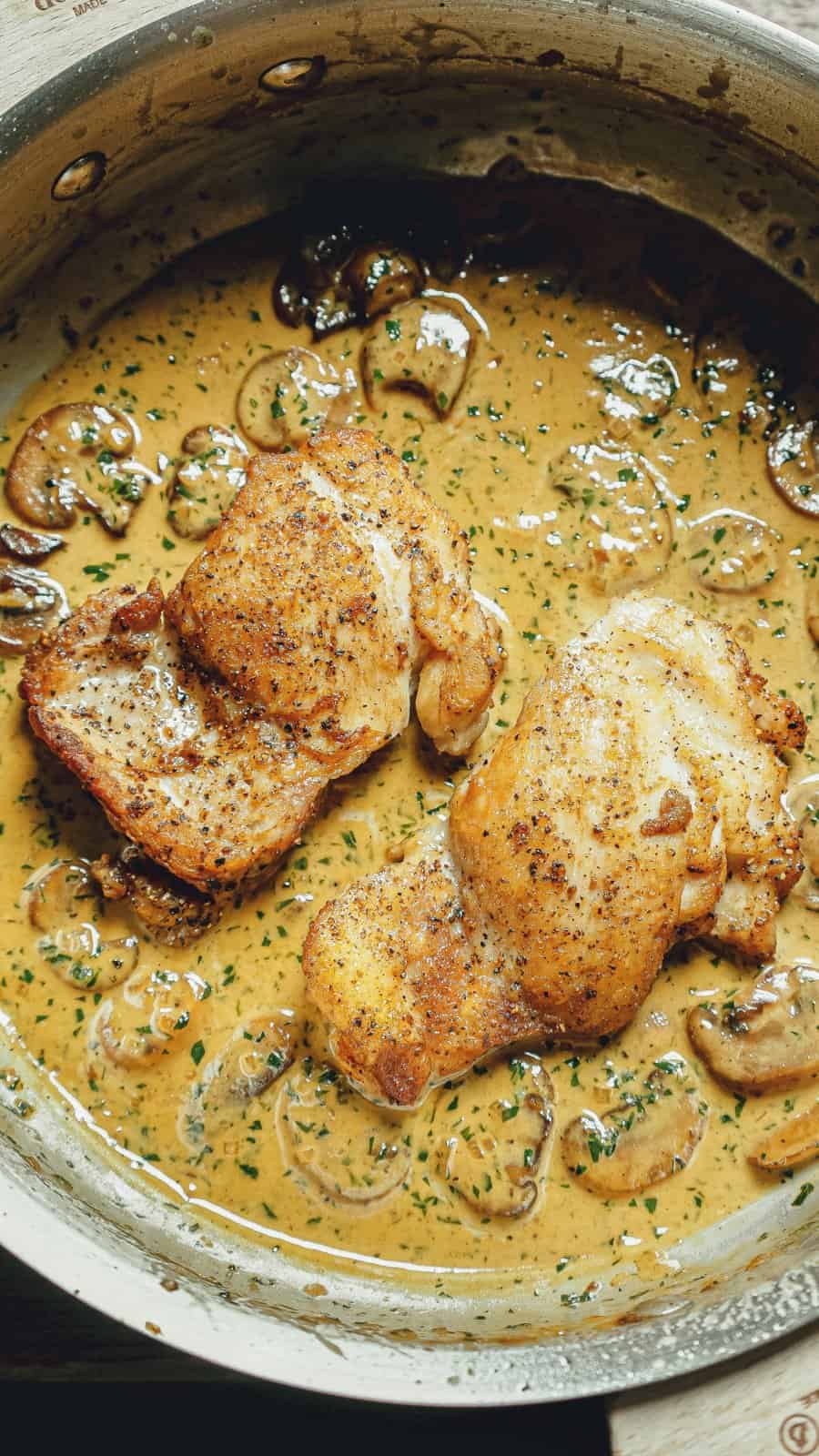 Two pieces of seasoned, golden-brown chicken in a creamy mushroom sauce with herbs, served in a stainless steel skillet.