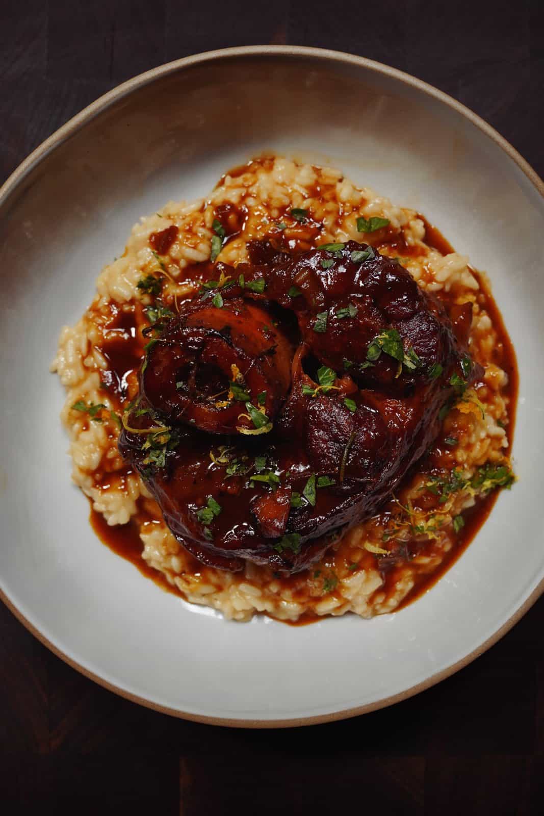 A bowl of creamy risotto topped with a glazed, braised meat shank, garnished with chopped herbs and surrounded by a rich brown sauce.