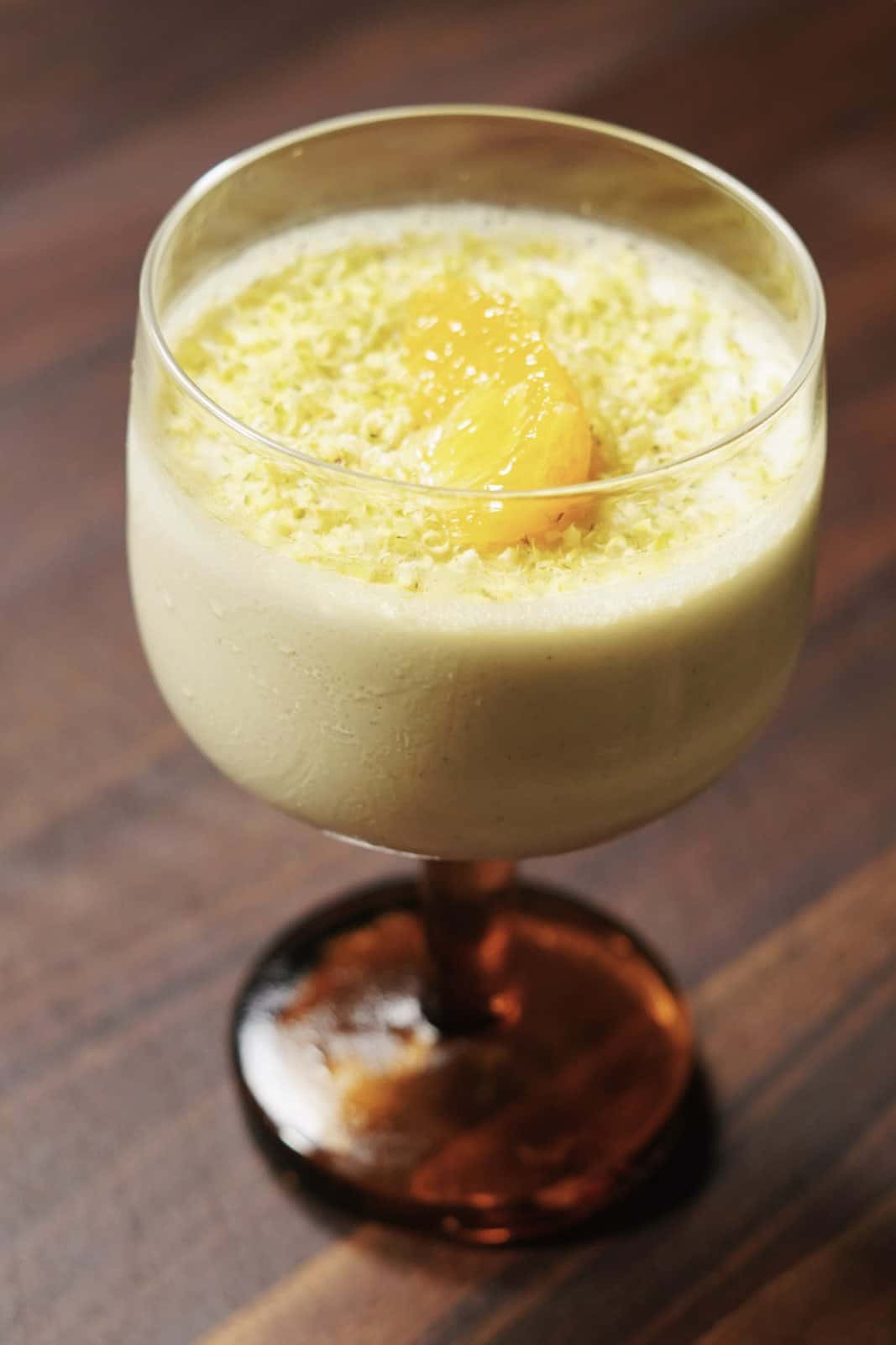 A dessert in a glass goblet with a creamy, light yellow base, topped with yellow zest and a dollop of fruit compote, set on a dark wooden surface.