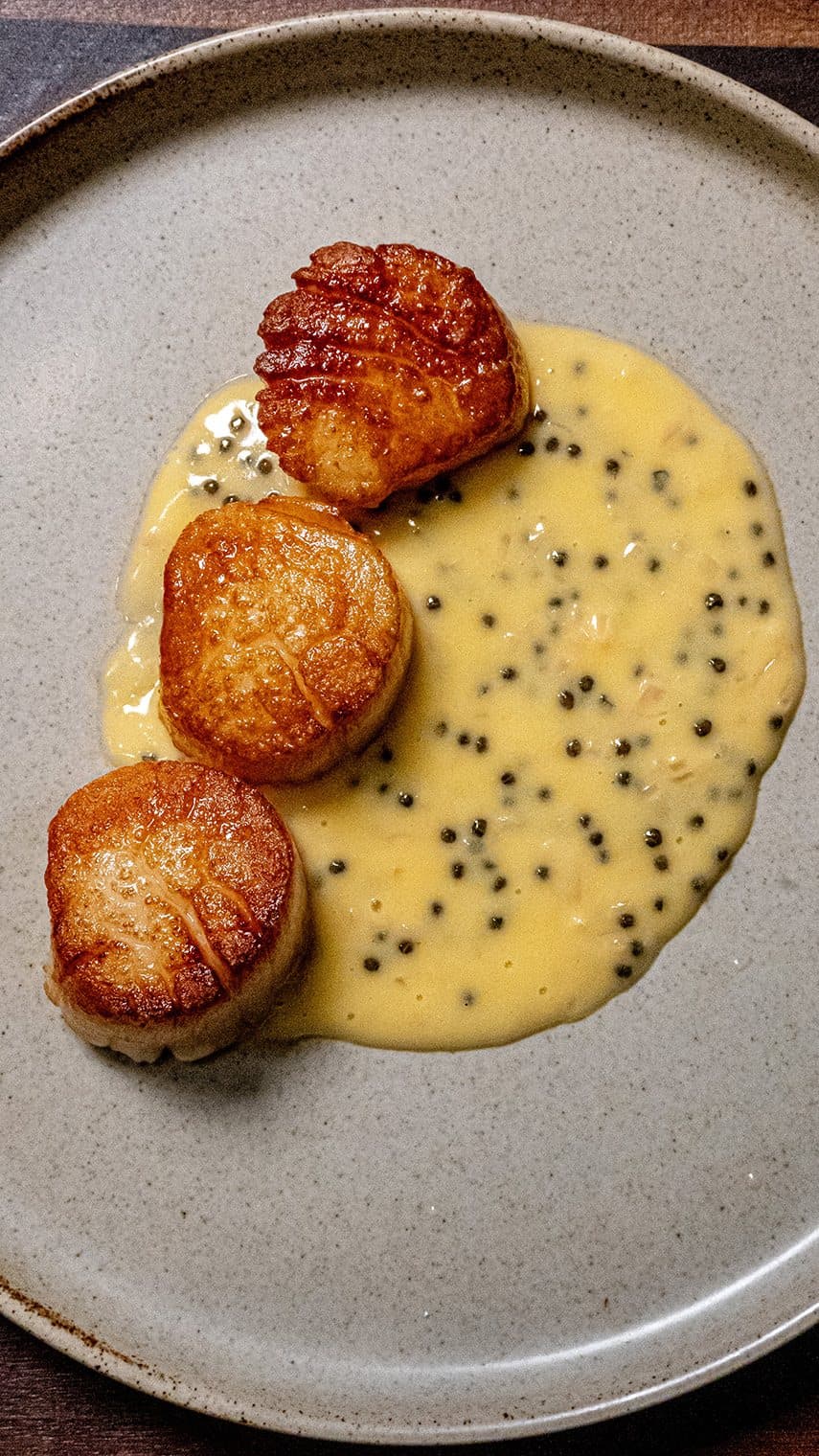 Three seared scallops are arranged on a plate, served atop a creamy yellow sauce with black specks, possibly caviar or pepper, on a light gray ceramic dish.
