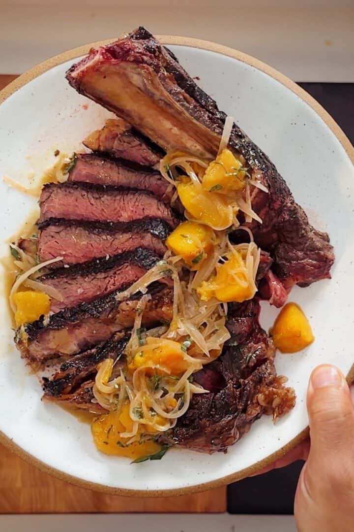 Overhead of a sliced bone-in chuletón ribeye on a white plate, fanned and topped with peach-and-shallot sauce, held over a wood board.