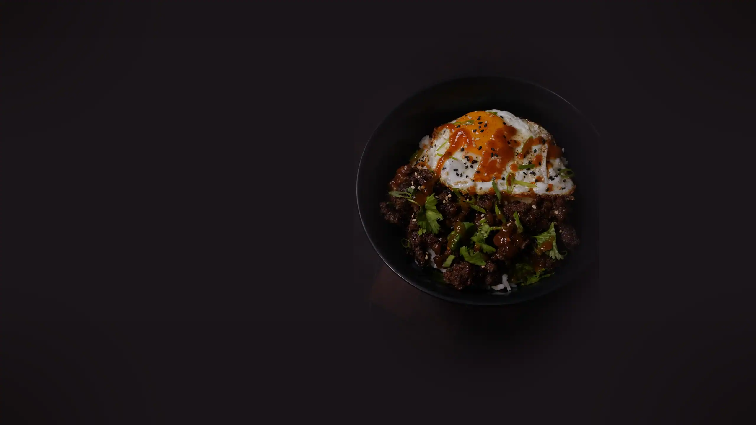 A rice bowl with seared beef, soft egg, and fresh herbs in a dark walnut bowl on an extended dark ground.