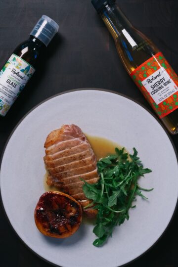 A plate with sliced cooked duck breast, arugula, and a grilled half peach, with bottles of balsamic glaze and sherry cooking wine nearby on a dark surface.