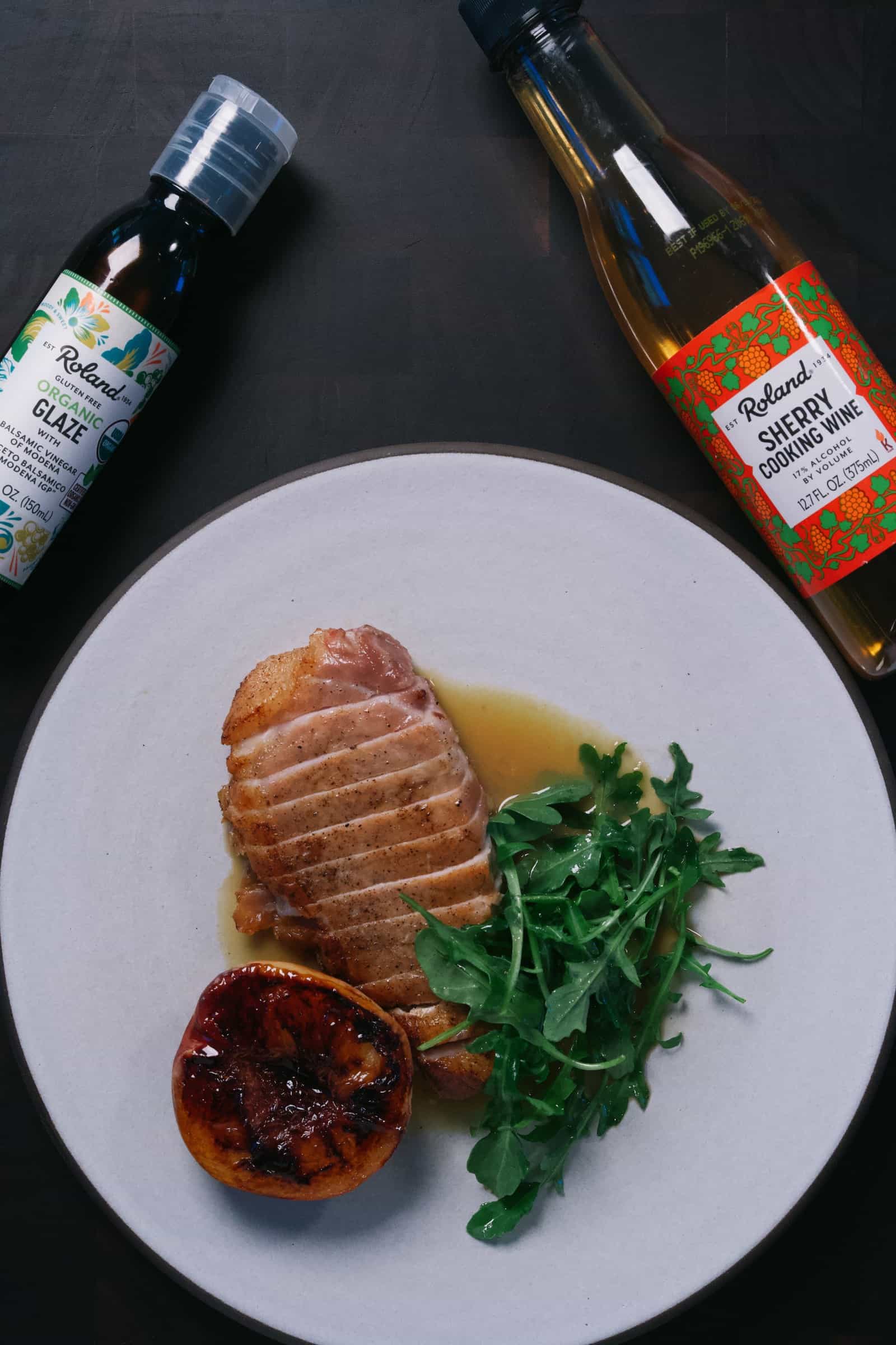 A plate with sliced cooked duck breast, arugula, and a grilled half peach, with bottles of balsamic glaze and sherry cooking wine nearby on a dark surface.