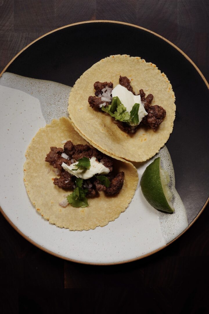 Two corn tortilla tacos filled with spiced ground beef, diced onion, cilantro and crema, with a lime wedge