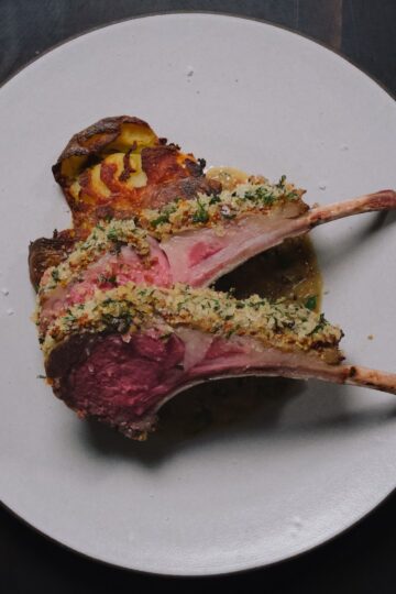 Two lamb chops sliced from a rack, a herb and mustard crust along the top edge, rare in the middle, on a white plate with pan sauce and a crisp smashed potato.