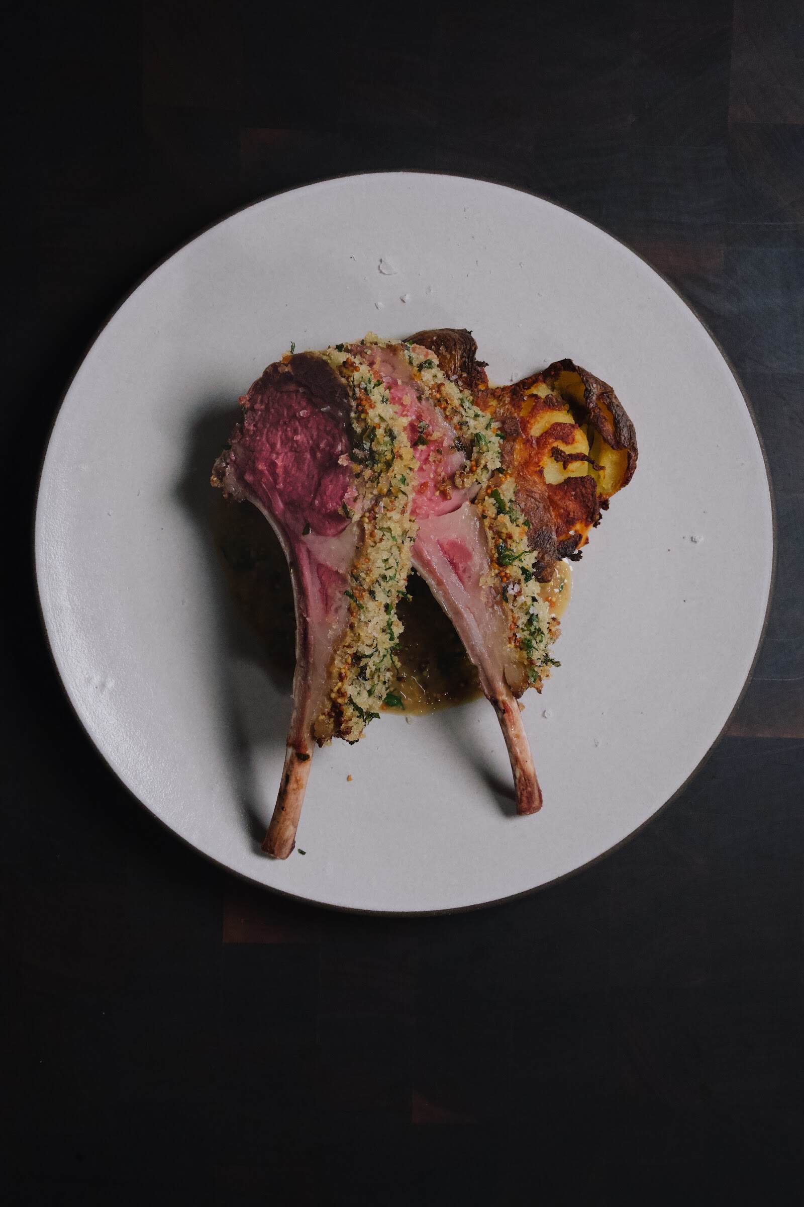 Two lamb chops standing on the bone, herb and mustard crust along the cut edge, rare in the middle, pan sauce and a smashed potato on a white plate.
