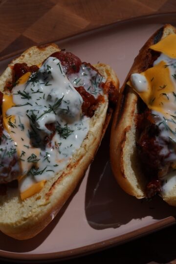 Two brioche buns holding venison boudin blanc sausages under venison chili, melted cheese, creme fraiche and chopped dill, on a pale oval plate over an end-grain butcher block.