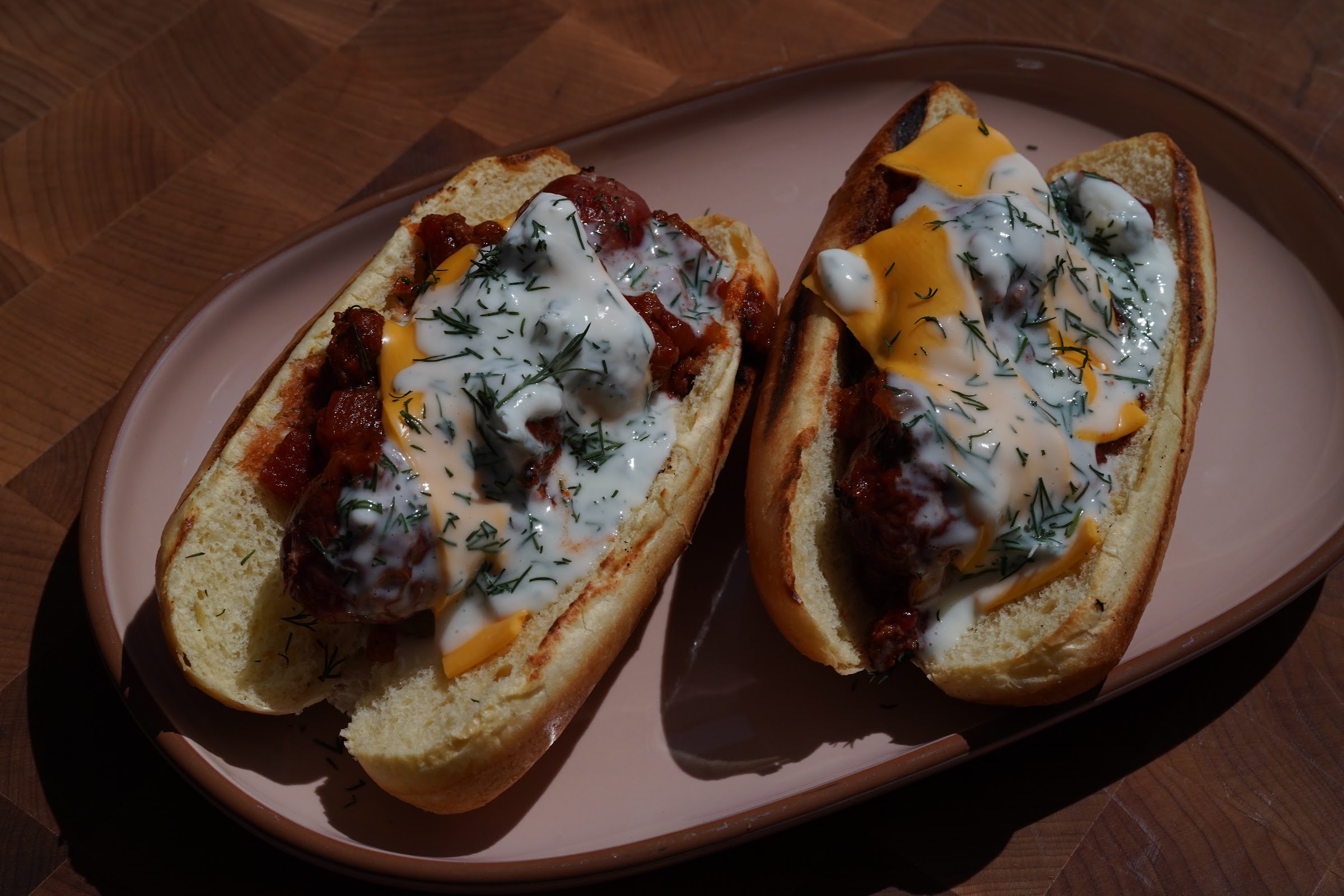 Two brioche buns holding venison boudin blanc sausages under venison chili, melted cheese, creme fraiche and chopped dill, on a pale oval plate over an end-grain butcher block.