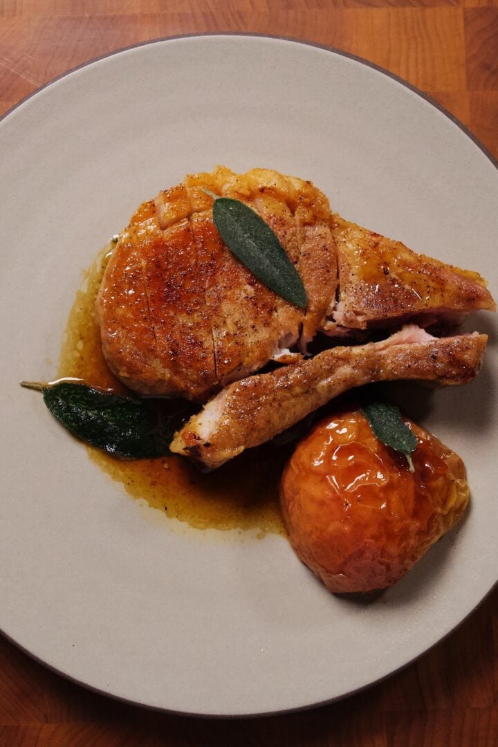 Overhead view of a cut, pan-seared pork chop on a light gray plate with a glazed roasted apple and sage leaves, on a butcher-block surface.
