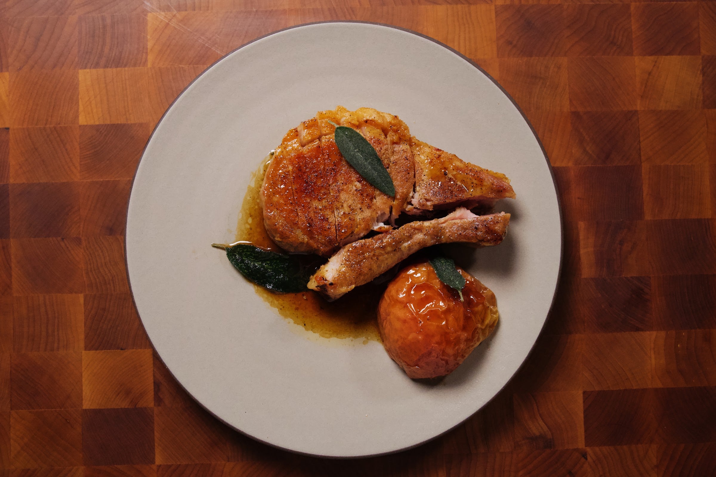 Overhead view of a cut, pan-seared pork chop on a light gray plate with a glazed roasted apple and sage leaves, on a butcher-block surface.